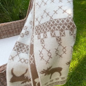 May include: A white and brown wool throw blanket with a Scandinavian-inspired design featuring moose and geometric patterns. The blanket is folded and draped over a wicker chair.