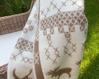 May include: A white and brown wool throw blanket with a Scandinavian-inspired design featuring moose and geometric patterns. The blanket is folded and draped over a wicker chair.