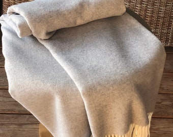 Puede incluir: Una manta suave de color gris con flecos blancos. La manta está doblada y colocada sobre una silla de mimbre.