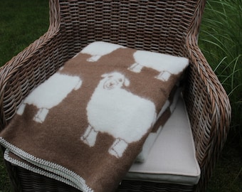 May include: A brown throw blanket with a white sheep pattern is folded and placed on a wicker chair. The chair is outdoors on a grassy surface.