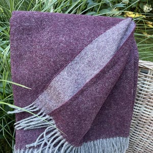 May include: A folded, plum-colored throw blanket with a lighter purple stripe and gray fringe. The blanket is made of a soft, textured material and is draped over a wicker chair, with greenery in the background.