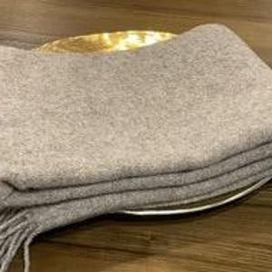 Puede incluir: Una manta suave de color marrón claro con flecos en los bordes. La manta está doblada y colocada sobre un plato dorado.