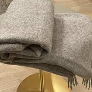 May include: A soft, grey throw blanket with a fringe border. The blanket is folded and displayed on a gold stand.