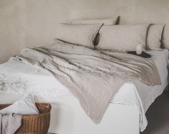 Puede incluir: Una cama blanca con un edredón de lino marrón claro y fundas de almohada a juego. La cama está hecha con una sábana blanca y tiene una cabecera de madera. Hay una pequeña cesta de mimbre con un paño blanco al pie de la cama.