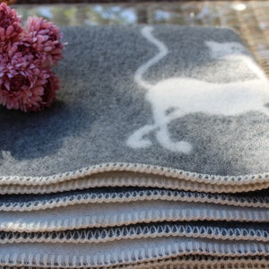 Puede incluir: Una manta de lana gris y blanca con un diseño de gato blanco. La manta está doblada y tiene un borde blanco de ganchillo.