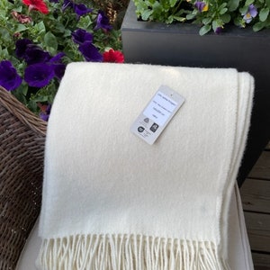 Peut inclure: Une couverture en laine blanche avec des franges sur le bord inférieur. La couverture est pliée en deux et porte une étiquette indiquant "100% laine de Nouvelle-Zélande, 130x170 cm".