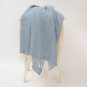 May include: A light blue throw blanket with a subtle diagonal weave pattern and white fringe trim along the bottom edges. The fabric has a soft, textured appearance, and the fringe consists of multiple thin, straight white strands of yarn.