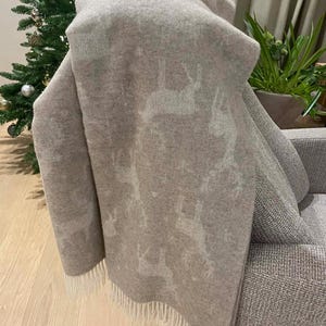May include: A beige throw blanket with a reindeer pattern draped over a grey sofa. The blanket has a fringe along the bottom edge. A Christmas tree with gold baubles is visible in the background.