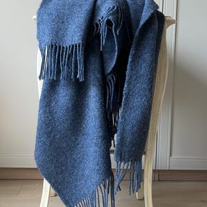 May include: A blue throw blanket with fringing draped over a white chair. The blanket has a soft, textured appearance and is a solid shade of blue. The fringing hangs from the bottom edge of the blanket.