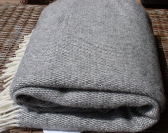 May include: A gray wool throw blanket with a woven texture and fringe on the edges. The blanket is folded and placed on a wicker surface.