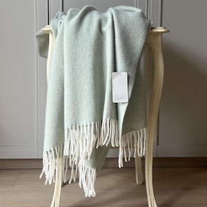 May include: A soft, light sage green throw blanket featuring a diagonal twill weave pattern and long, white fringe along the bottom edge. A small, rectangular white product tag is attached to the fabric, displaying minimal black text.