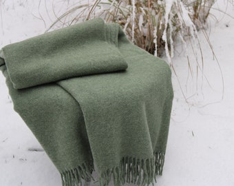 May include: A sage green blanket with a folded section on top. The blanket has a soft, textured appearance and a fringed edge. The blanket is draped over a surface, with a snowy background and some dried plants.