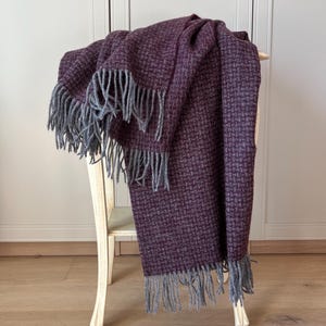 May include: A woven throw blanket featuring a deep purple and dark grey checkered pattern. The blanket has long, grey fringe detailing along the edges, providing a textured finish to the fabric. The textile displays a consistent grid design.