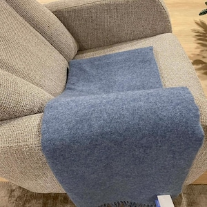 Könnte beinhalten: Eine blaue Decke mit Fransen, die über einem beigen Sessel drapiert ist. Die Decke hat ein weiches, strukturiertes Aussehen. Der Sessel ist mit einem gewebten Stoff bezogen. Die Szene deutet auf einen komfortablen Wohnraum hin.