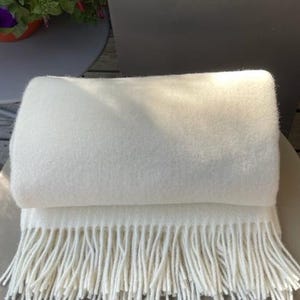 May include: A white throw blanket with a fringe border. The blanket is folded in half and is sitting on a white surface.