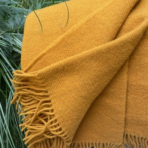 Puede incluir: Manta tejida de color amarillo mostaza con bordes con flecos. La manta está doblada, mostrando su textura y calidez. La franja añade un elemento decorativo al textil para el hogar acogedor.
