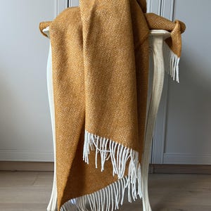 May include: A mustard yellow throw blanket featuring a classic herringbone pattern throughout the fabric. The blanket is finished with long, white twisted fringe tassels along the bottom edges, providing a distinct contrast to the warm yellow tone.