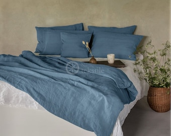 Puede incluir: Una funda nórdica de lino azul con una sábana de lino blanco debajo. La funda nórdica está doblada hacia atrás para revelar la sábana blanca. Hay cuatro almohadas de lino azul en la cama. La cama está contra una pared verde claro. El texto "Namu tekstilė" es visible en la cama.