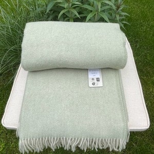 Peut inclure: Une couverture en laine douce de couleur vert clair avec un motif en chevrons subtil et une bordure à franges. La couverture est pliée et présentée sur une surface de couleur crème.