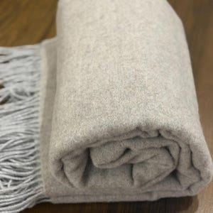 Può includere: Una coperta grigio chiaro arrotolata, con un bordo a frange. La coperta è realizzata in un materiale morbido e strutturato e poggia su una superficie di legno. La frangia è di una tonalità di grigio più chiara.