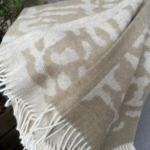 May include: A folded beige and white patterned throw blanket with a fringe. The blanket features a repeating pattern of abstract shapes in a lighter beige against a darker beige background. The fringe is cream-coloured.