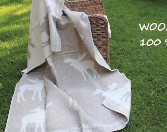 May include: A beige wool throw blanket with a white moose design. The blanket is folded and draped over a wicker chair. The text "WOOL 100%" is written on a white background.