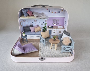 May include: A miniature dollhouse made from a suitcase with a pink exterior. The interior is decorated with a living room and kitchen. The living room has a gray couch, two armchairs, a coffee table, and a rug. The kitchen has a white countertop, a sink, and a stove. The dollhouse is decorated with rainbow wallpaper and a string of lights.