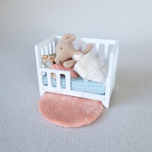 May include: White wooden doll's cot with a light blue and floral patterned mattress, a light brown plush toy animal lying on top of the mattress, and a white blanket. The cot is sitting on a light pink round rug.