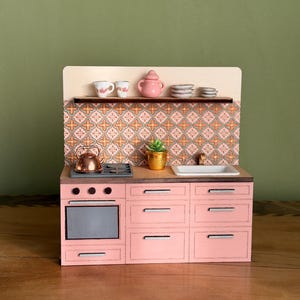 Könnte beinhalten: Eine Miniatur-Küchenzeile in Rosa mit Herd, Backofen, Spüle und Schränken. Das Set enthält einen Kupferkessel, einen kleinen goldenen Topf und ein Regal mit Geschirr und einer Teekanne. Der Fliesenspiegel hat ein Muster.