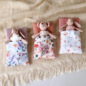 May include: Three stuffed animal toys are lying in their beds. Each bed is a small pillow with a floral print fabric cover. The toys are a brown bunny, a brown bear, and a white bunny. The beds are on a beige fabric background.