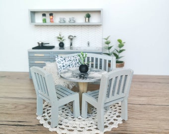 May include: A miniature dining set with a round table and four chairs. The table has a white marble top and a grey base. The chairs are also grey and have a slatted back. The set is on a white crocheted doily. There is a small plant in a black pot on the table.