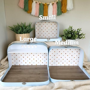 May include: Three light blue vintage-style suitcases in small, medium, and large sizes. The interiors feature a polka dot pattern and a wooden base. The small suitcase is closed, while the medium and large are open, showcasing the interior design. The word "Small", "Large", and "Medium" are written above the suitcases.