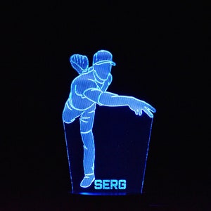 Pode incluir: Uma luz noturna impressa em 3D azul que representa um jogador de beisebol em uma pose de arremesso. A base da luz tem o nome "SERG" impresso.