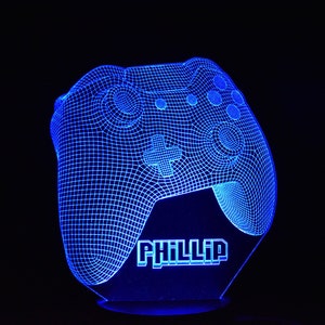 Può includere: Lampada a LED blu a forma di controller di gioco, con il nome "PHILLIP" illuminato alla base. Il design del controller presenta un motivo a griglia, pulsanti e un D-pad. Lo sfondo è nero.