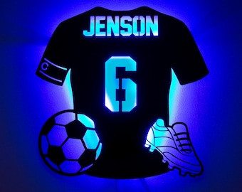 May include: Black soccer jersey-shaped wall art with the name "JENSON" and the number "6" illuminated in blue. Includes a soccer ball and a soccer cleat, also with blue illumination. A decorative piece for a sports enthusiast.