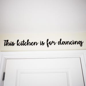 May include: Black cursive text on a cream-coloured wall reads "This kitchen is for dancing." The text is above a white door. The image is well-lit and suitable for kitchen decor.