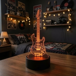 May include: An orange LED light shaped like an electric guitar with the name TOM printed at the base. The guitar outline is etched onto a clear acrylic panel that sits on a solid black cylindrical base, emitting a warm glow in a dark room.