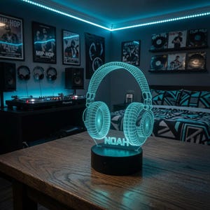 Op de afbeelding: Een 3D-stijl LED-lamp met een oplichtende cyaankleurige draadmodelafbeelding van een hoofdtelefoon. De voet van de lamp is zwart en cilindervormig, met de naam NOAH in cyaankleurige blokletters in het midden van het hoofdtelefoonframe.
