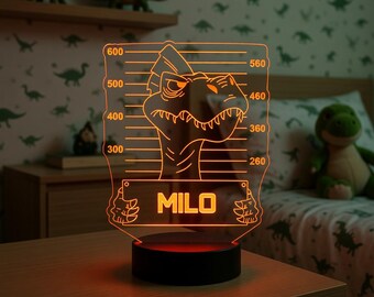May include: A glowing orange dinosaur-themed night light with the name "MILO" displayed on a rectangular base. The light features a dinosaur design with height markings. The base is black, and the background shows a dinosaur-themed bedroom.
