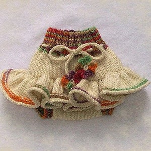 Puede incluir: Una falda de bebé tejida con base crema y ribete de colores. La falda tiene un diseño con volantes y un lazo en la cintura.