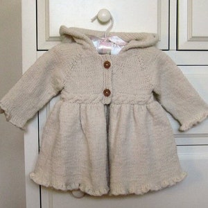 May include: A cream-colored knitted baby cardigan with a hood and a button closure. The cardigan has a ruffled hem and cuffs.