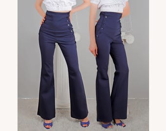 May include: Navy blue high-waisted pants with a flared leg. The pants feature a row of buttons on each side of the waist. The model is wearing blue open-toe heels. The pants are a classic style.
