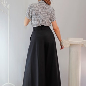 May include: A woman wearing a short-sleeved blouse with a small geometric pattern in black and white, paired with wide-leg black pants. The pants have a high waist and a flowing silhouette. The outfit is simple and elegant.