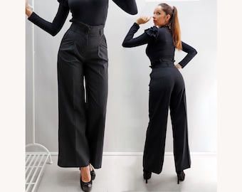 May include: High-waisted black trousers featuring a subtle vertical pinstripe pattern, a wide-leg silhouette, and a structured waistband with belt loops. The trousers are paired with a long-sleeved black top with ruffled shoulder detailing.