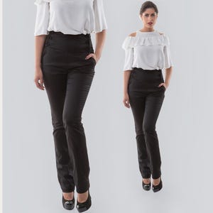 May include: High-waisted black trousers featuring a side-laced detail at the waistband, straight-leg cut, and side pockets. The pants are paired with a white short-sleeved top with a ruffled off-the-shoulder neckline and black high-heeled shoes.