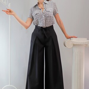 May include: A woman wearing a short-sleeved button-down shirt with a small, repeating pattern in black and white, paired with high-waisted, wide-leg black pants. The pants have a tie at the waist. The outfit is completed with black sandals.