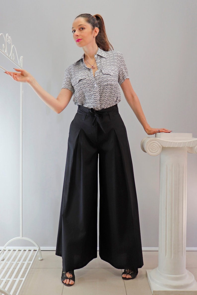 May include: A woman wearing a short-sleeved button-down shirt with a small, repeating pattern in black and white, paired with high-waisted, wide-leg black pants. The pants have a tie at the waist. The outfit is completed with black sandals.