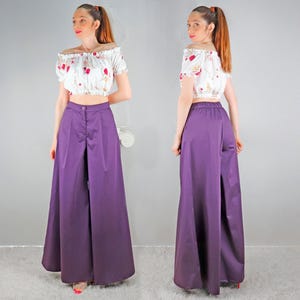 May include: A two-piece outfit featuring a white off-the-shoulder crop top with a floral pattern and high-waisted, wide-leg purple pants. The pants have a button closure and a split detail in the front. The outfit is suitable for a casual or semi-formal occasion.