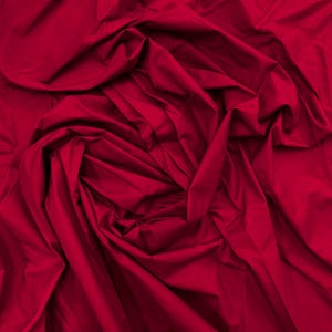 May include: A solid deep red fabric with a matte finish, featuring numerous folds and creases that create a dynamic, layered texture across the entire surface. The material appears soft and opaque, with shadows highlighting the depth of the fabric.