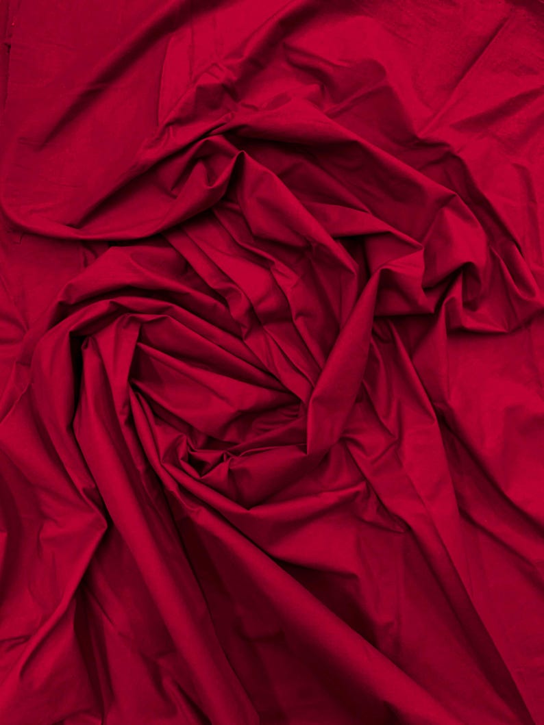 May include: A solid deep red fabric with a matte finish, featuring numerous folds and creases that create a dynamic, layered texture across the entire surface. The material appears soft and opaque, with shadows highlighting the depth of the fabric.